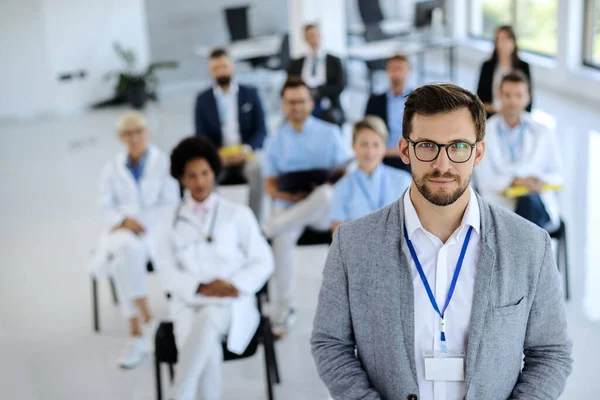 Young businessman leading a seminar to group of healthcare workers and business people in board room and looking at camera.