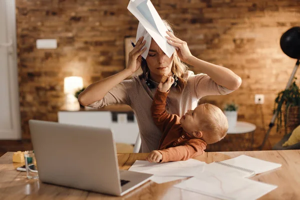 Stressed working mother covering her head with paperwork while her small son is offering her a cookie at home.