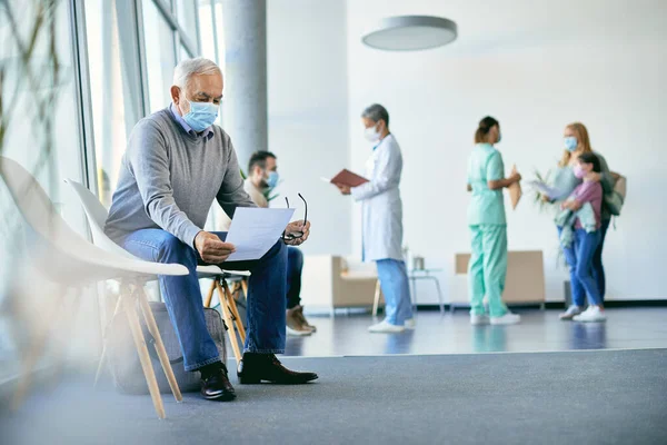 Senior man with face mask analyzing medical documents while sitting in waiting room after medical exam at the hospital.