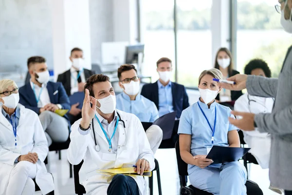 Happy male doctor wearing face mask and raising his hand to ask something on a seminar at conference hall.