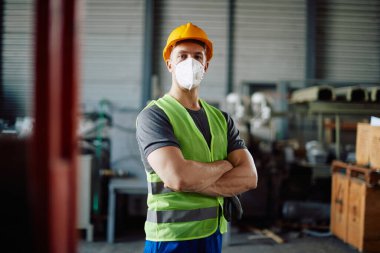 Warehouse worker standing with arms crossed at storage compartment and wearing face mask due to COVID-19 pandemic.