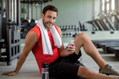 Young happy athlete taking a break from exercising and texting on cell phone at health club. 