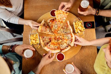 High angle view of unrecognizable friends sharing pizza while drinking beer in a pub. 