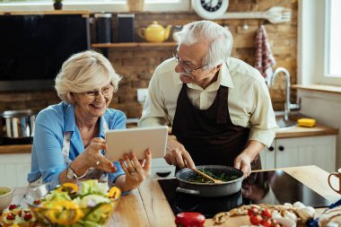 Happy senior couple using digital tablet and having fun while preparing lunch in the kitchen. 