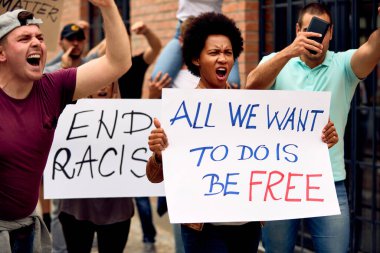Multi-ethnic crowd of people marching for racial equality on city streets. Focus is on African American woman shouting while holding a placard with freedom inscription. 