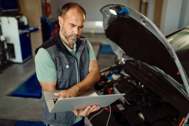 Auto mechanic using laptop while examining engine performance of a car at workshop. 