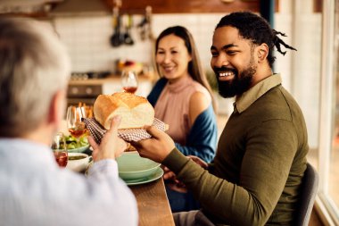 Happy African American man having lunch with friends and passing bread to one of them at dining table. 