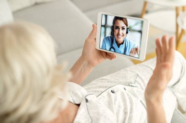 Close-up of mature woman talking online with her doctor while using digital tablet at home. Focus is on female doctor on touchscreen. 