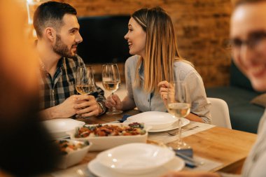 Young happy couple talking to each other while drinking wine and having dinner with friends at dining table. 