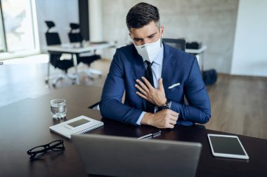 Male entrepreneur with protective face mask working on a computer and feeling chest pain in the office.