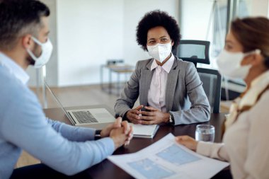 African American real estate agent talking to a couple while going through blueprints during the meeting. They are wearing protective face masks due to coronavirus pandemic. 