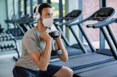 Male athlete using hand weight while doing squats and wearing protective face mask in health club. 