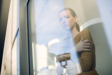 Low angle view of young sad woman looking through the window and thinking of something. 