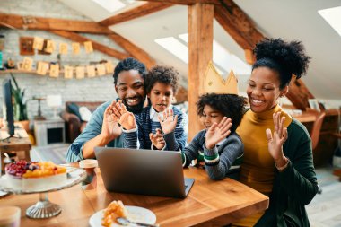 Happy African American family celebrating daughter's Birthday and waving to someone during video call over laptop at home.