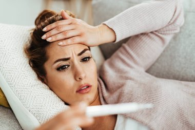Worried woman measuring her body temperature with thermometer while lying down on sofa at home. 