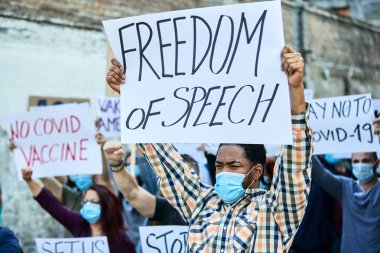 African American man wearing protective face mask while participating in public demonstrations and carrying banner with 'freedom of speech' inscription. 