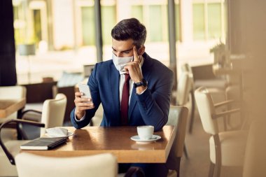 Pensive businessman wearing protective face mask while text messaging on mobile phone in a cafe. 