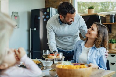 Happy man serving food and talking to his wife while having family lunch at dining table. 