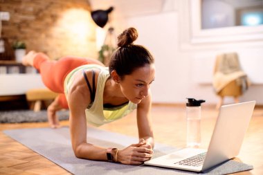 Dedicated sportswoman working out while following online exercise class over laptop at home.