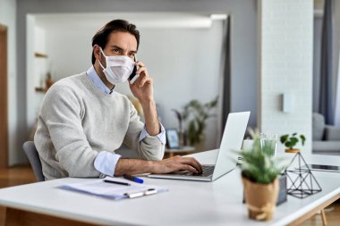Young businessman wearing protective face mask while working on laptop and talking on mobile phone at home office. 