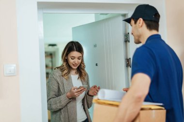 Young woman text messaging on mobile phone while standing on a doorway and receiving package from delivery man. 