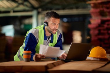 Male worker talking via headset while using laptop and going through delivery plans at distribution warehouse.