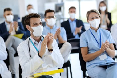 Male doctor and his colleagues wearing protective face masks and clapping their hands after successful education event in convention center. 