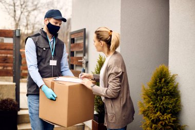 Young woman signing for home delivery and communicating with courier who is wearing protective face mask due to coronavirus pandemic. 