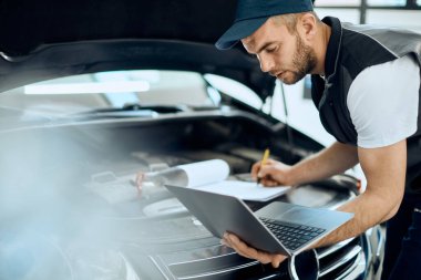 Young repairman using computer and taking notes while examining car engine in a workshop.