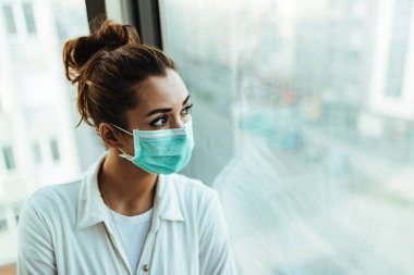 Young distraught woman with face mask standing by the window and looking through it. 