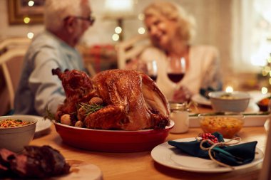 Traditional roast turkey at dining table during Thanksgiving meal at home. Senior couple is in the background. 