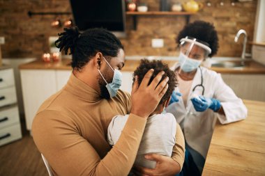 Rear view of a small black boy sitting in father's  lap while receiving vaccine due to coronavirus pandemic. 