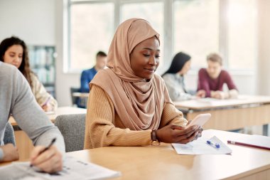 Smiling black Islamic student texting on cell phone during a class at college classroom. 