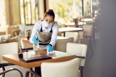 Waitress cleaning tables with disinfectant and wearing protective face mask in a cafe during COVID-19 pandemic.