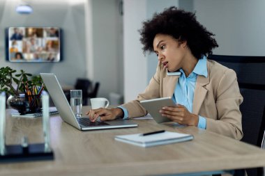 Ofiste telefonla konuşurken laptopta ve dokunmatik ekranda çalışan çok görevli siyahi iş kadını.. 