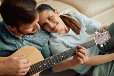 Smiling woman enjoying while her husband is playing acoustic guitar for her at home.