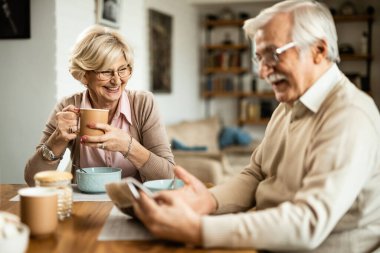 Happy mature couple eating breakfast and reading newspaper in the morning at dining table. Focus is on happy woman. 