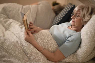 Senior woman relaxing while reading a book in bed in the evening. Her husband is sleeping in the background. 