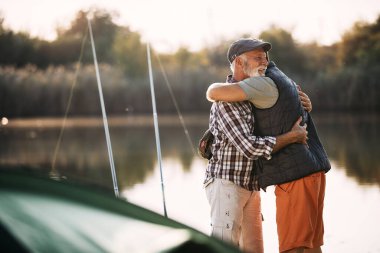 Happy mature man embracing his son while camping and fishing on the lake. Copy space.
