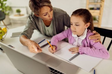 Little girl learning at home with help of her mother who is explaining her assignments on laptop. 