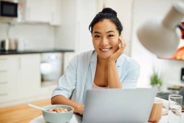 Young happy Asian woman using laptop while having breakfast at home and looking at camera. 