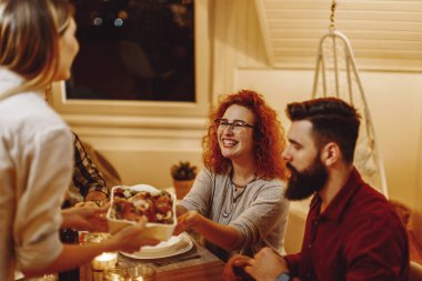 Group of happy young people having dinner together at dining table. Focus is on redhead woman taking food from her friend. 