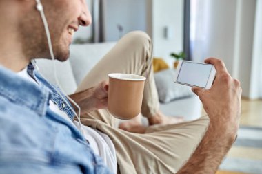 Close-up of  man drinking coffee while using smart phone and relaxing in the living room. Focus is on blank device screen. Copy space. 