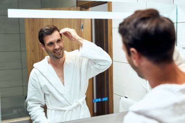 Happy man in bathrobe looking him self in a mirror in the bathroom. 