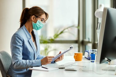 Young businesswoman wearing protective face mask while going through reports and working in the office. 