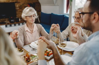 Extended family saying grace while holding hands at dining table. Focus is on senior woman. 