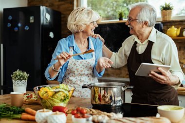Happy senior couple talking while using digital tablet and making lunch in the kitchen.