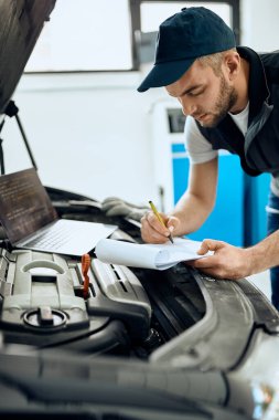 Auto repairman writing data while running diagnostic of car engine in a workshop. 