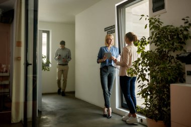 Happy businesswomen communicating while standing in hallway of an office building. Focus is on businesswoman holding digital tablet.