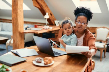 African American little girl using cell phone and sitting on mother's lap while she going through paperwork and working on laptop at home.